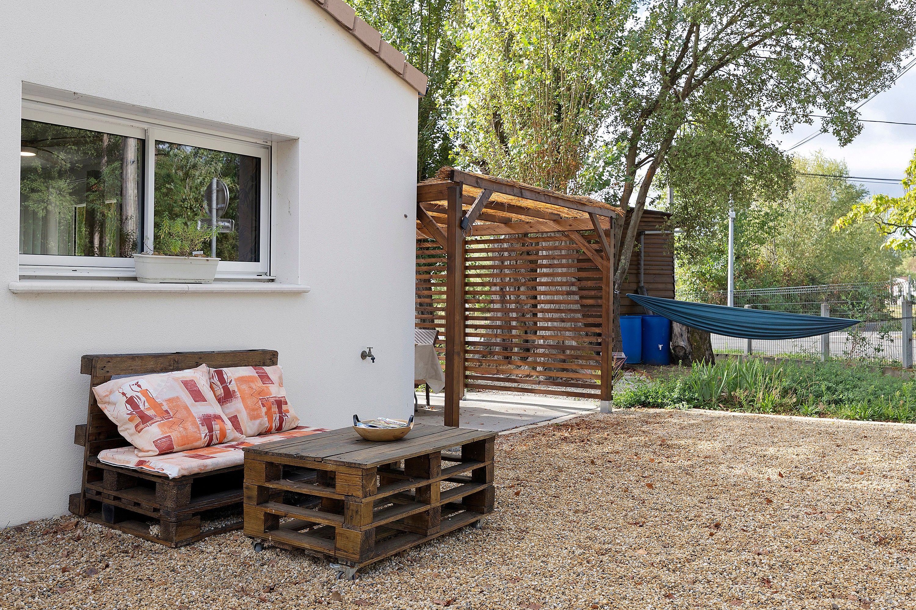 Un área de estar al aire libre de estilo rústico con un banco de madera y una mesa, una pérgola de madera y una hamaca.