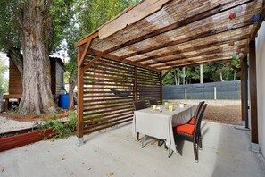 En uteplats med en träpergola, ett bord för två personer med stolar, och ett stort träd i bakgrunden.