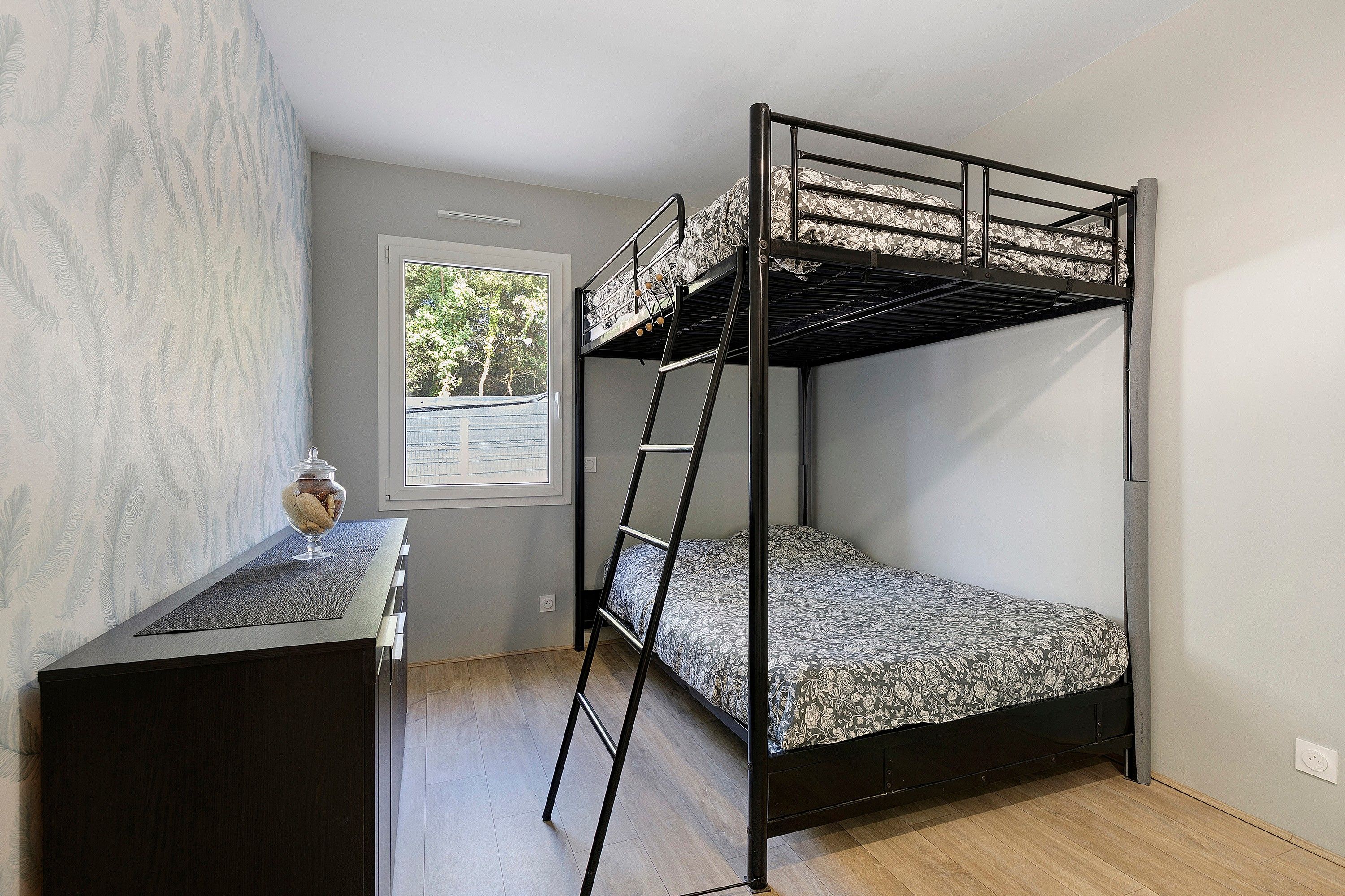 A bunk bed with a ladder, a small table with a glass container, and a window with a view of trees.