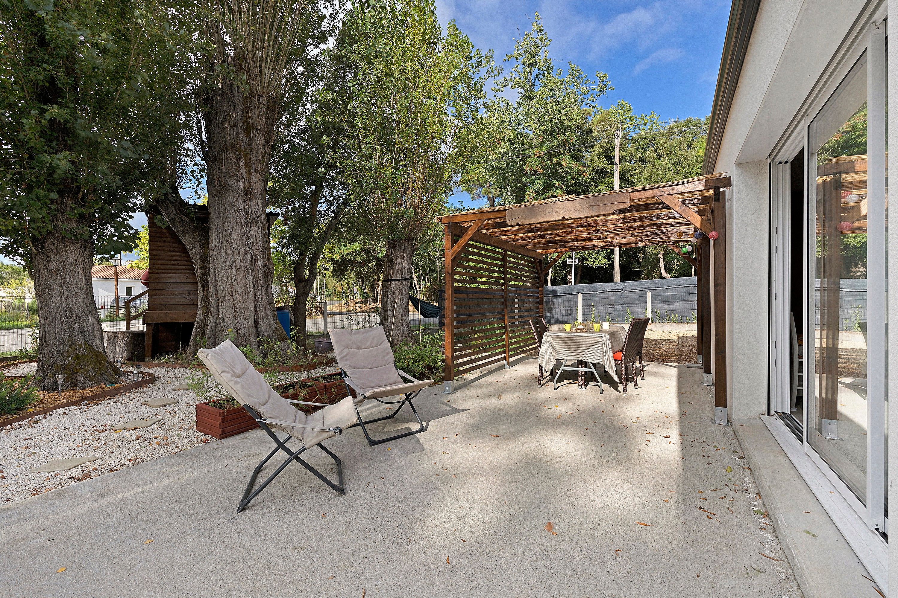 Un patio exterior con una pérgola, dos sillones, una mesa con sillas y un árbol grande.