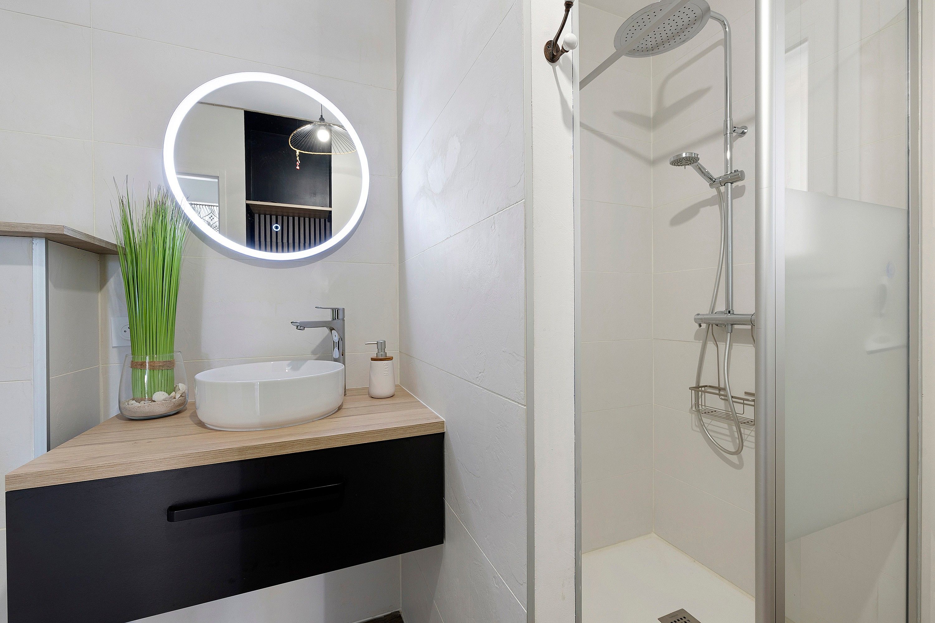 Une salle de bain moderne avec un miroir rond, un lavabo, une douche équipée d’un pommeau de douche à main et un espace de douche délimité par des parois en verre.
