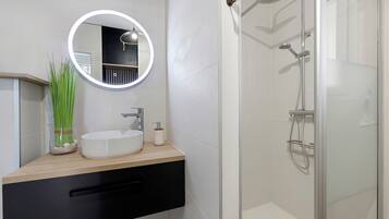 Une salle de bain moderne avec un miroir rond, un lavabo, une douche équipée d’un pommeau de douche à main et un espace de douche délimité par des parois en verre.