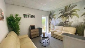 A living room with a sofa, a coffee table, a plant, a cabinet, and a mural of palm trees. - Charming house in the heart of L'Aiguillon-sur-mer - 8 people (L'Aiguillon-la-Presqu'île)