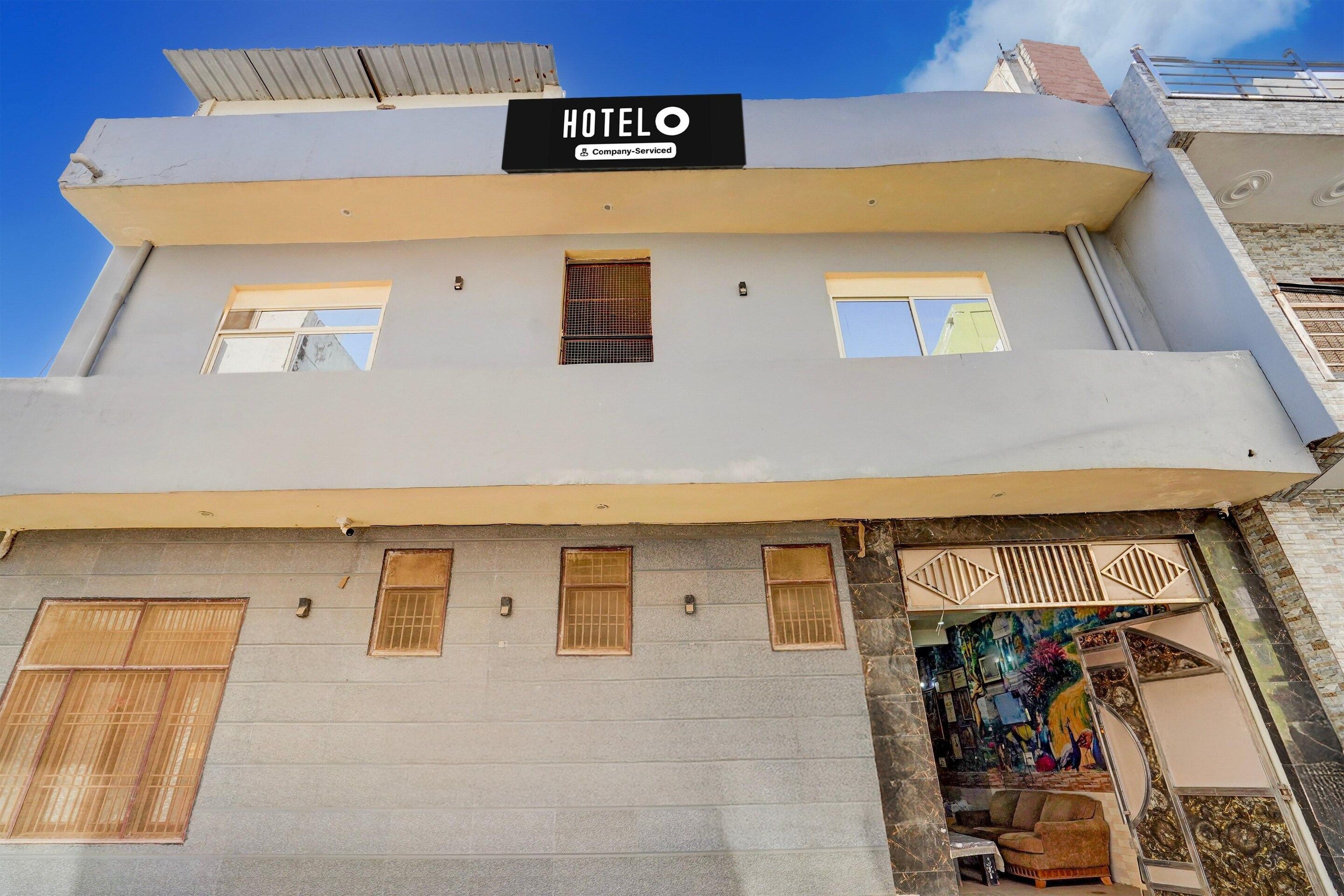 A hotel building with a sign indicating it is company-serviced. There are multiple windows on the facade, and a small outdoor area with a colorful mural.