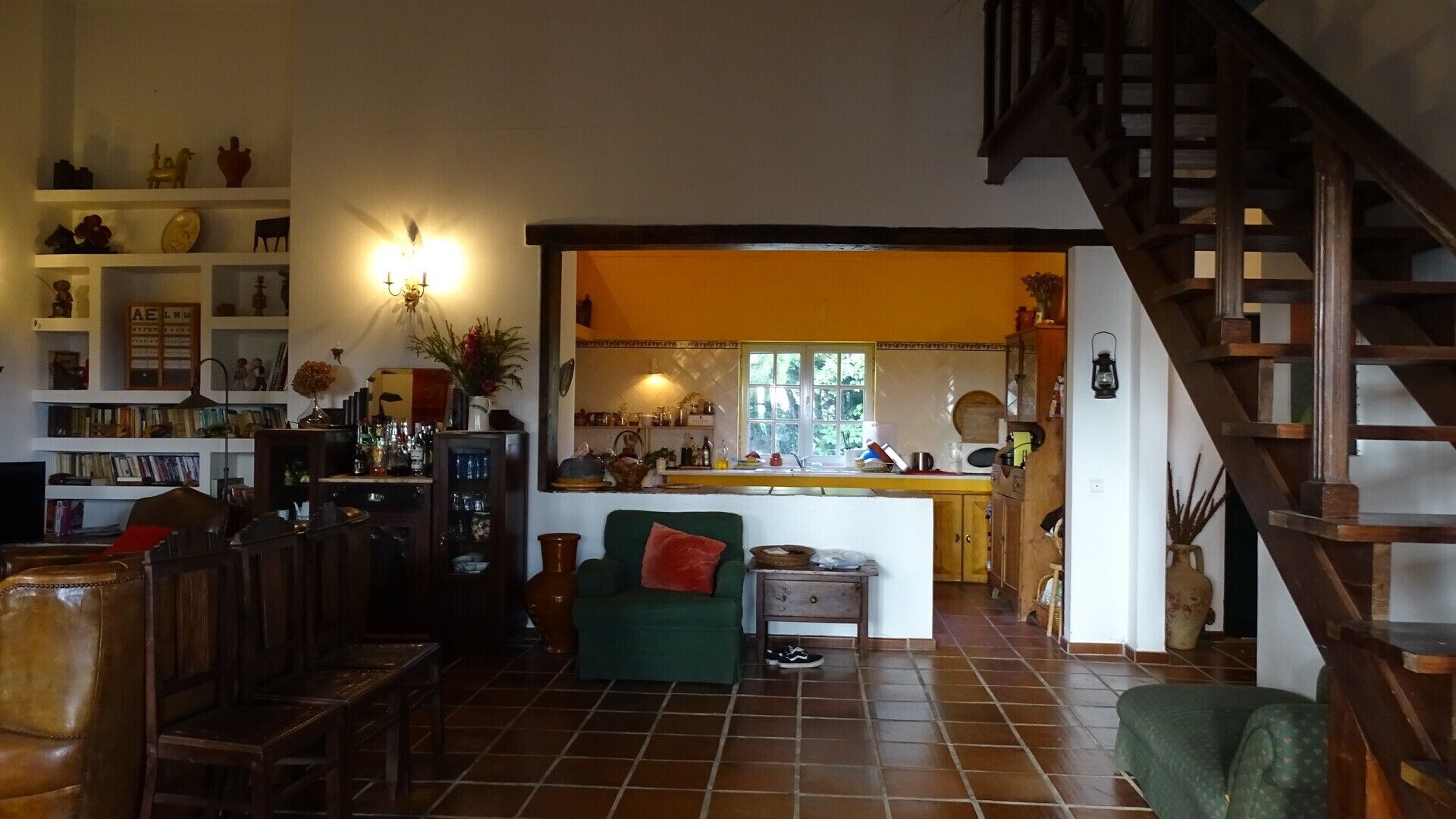 A cozy living room with a green sofa, a wooden staircase, and a kitchen area with a window.