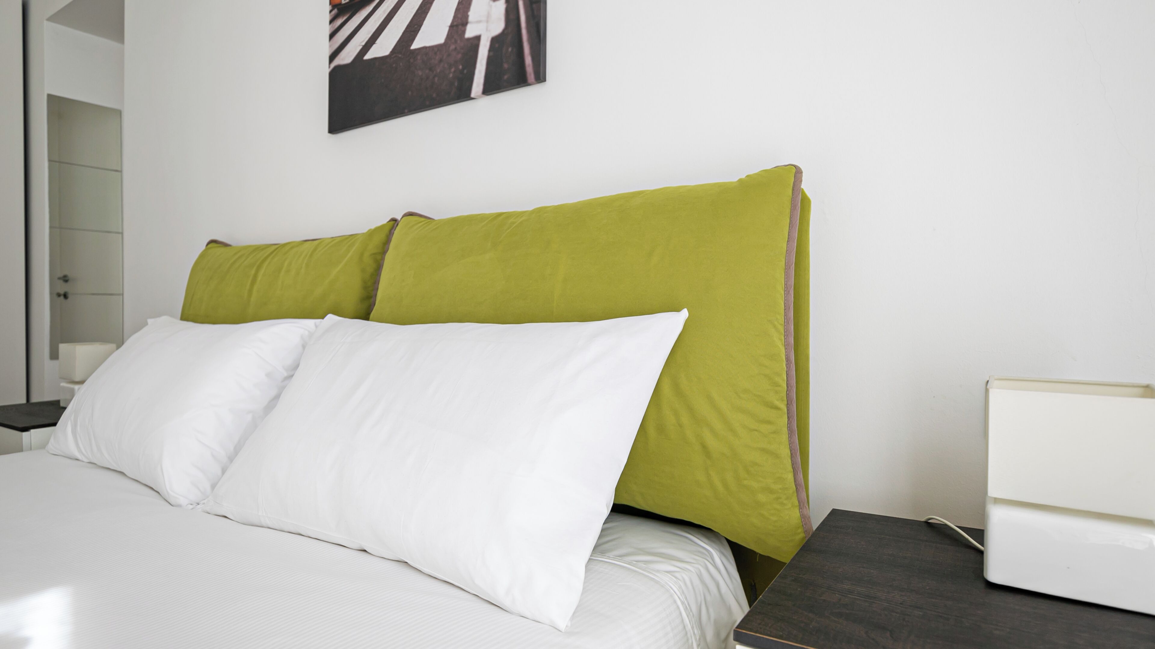 A hotel room with a bed, white pillows, a green accent pillow, a white nightstand, and a white bedside table.