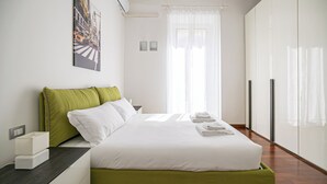 Ett hotellrum med en säng, gröna kuddar, vita sängkläder, ett nattduksbord, en inramad tavla, ett fönster med tunna gardiner och en garderob.