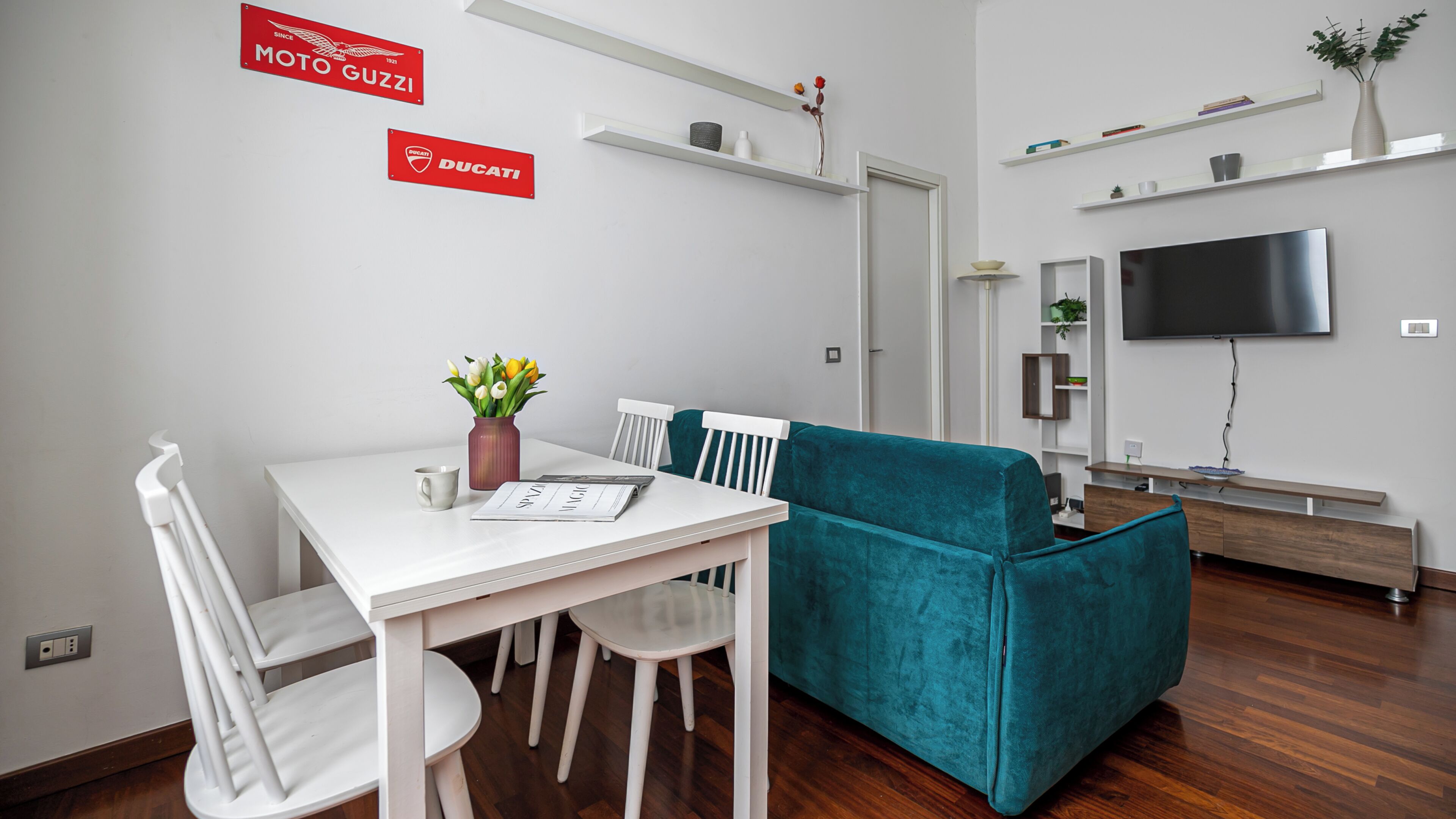 A modern living room with a teal sofa, a white dining table with chairs, a TV mounted on the wall, and a mounted shelf with decorative items.