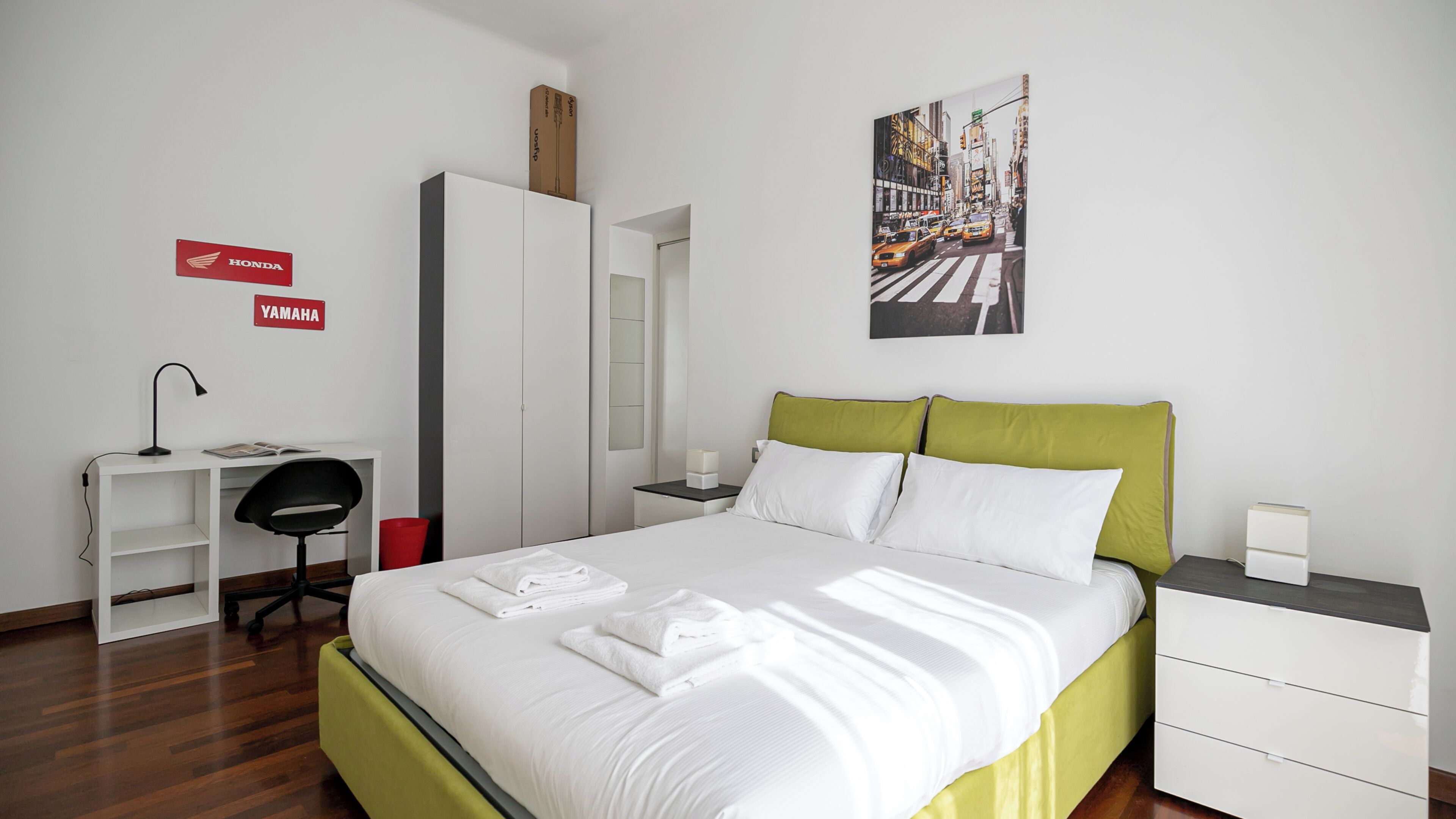 Camera d'albergo con un letto, una scrivania, una sedia, un armadio e un dipinto che raffigura un panorama urbano.