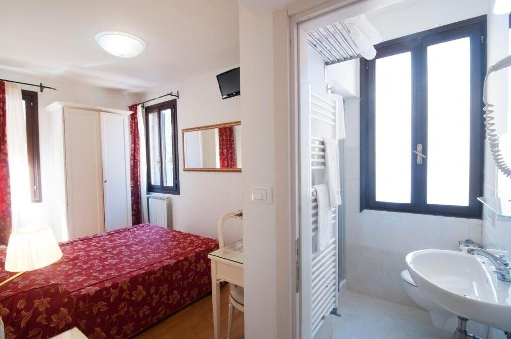 A bedroom with a bed, a chair, a desk, a mirror, a window, and a bathroom with a sink.