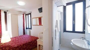 A bedroom with a bed, a chair, a desk, a mirror, a window, and a bathroom with a sink.
