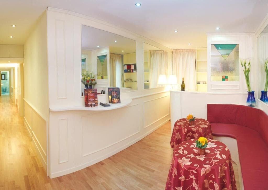 A hotel room with a curved white console, a red sofa, a small table with a floral tablecloth, and a large mirror reflecting the room.