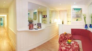 A hotel room with a curved white console, a red sofa, a small table with a floral tablecloth, and a large mirror reflecting the room.