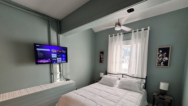 Un dormitorio con cama, televisor montado en la pared, ventilador de techo y ventana con cortinas.