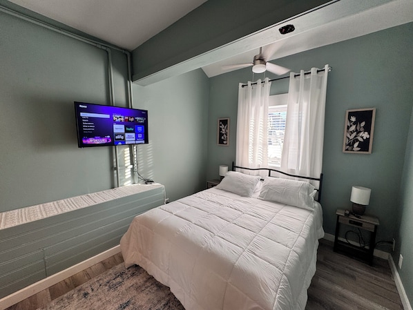 A bedroom with a bed, a television mounted on the wall, a ceiling fan, and a window with curtains.