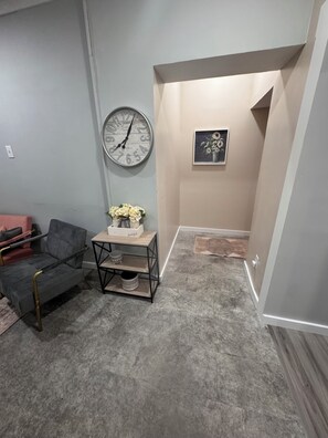 A room with a grey couch, a small table with a vase, a clock, and a framed picture on the wall.