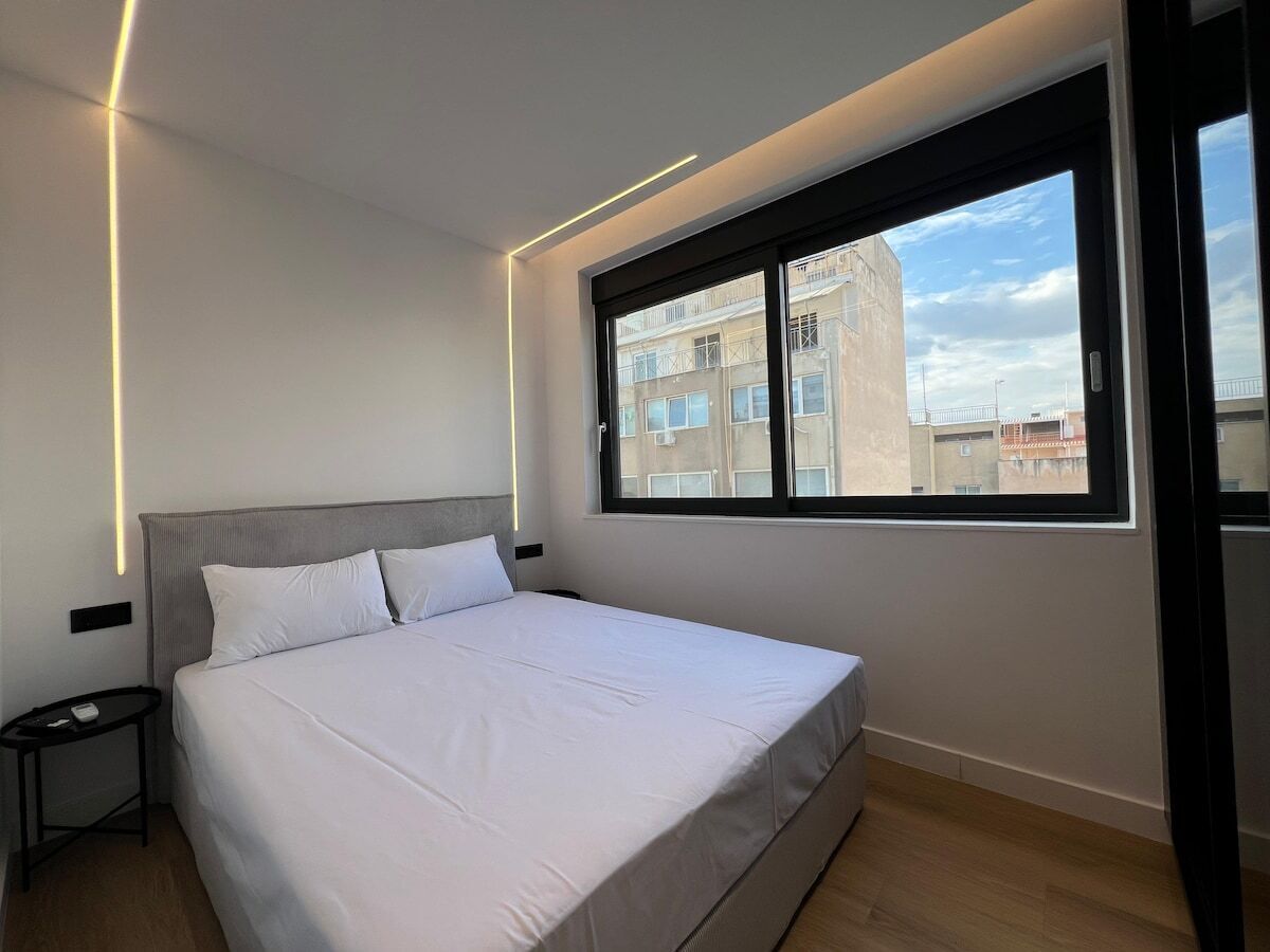 A modern hotel room with a large bed, a bedside table, a window with a view of buildings, and a ceiling light.