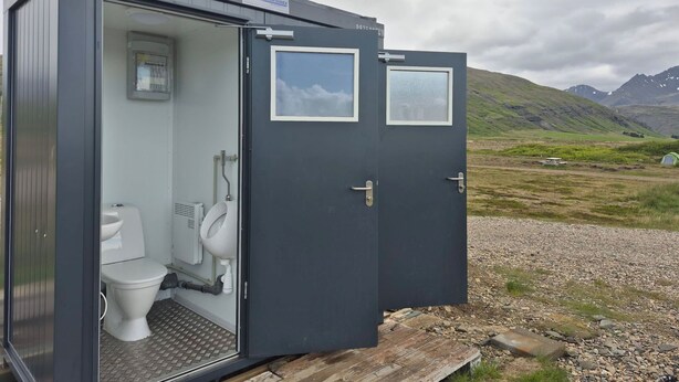 A portable restroom with a toilet and urinal, a window, and a door. - Stafafell Nature Park Camping (Stafafell)