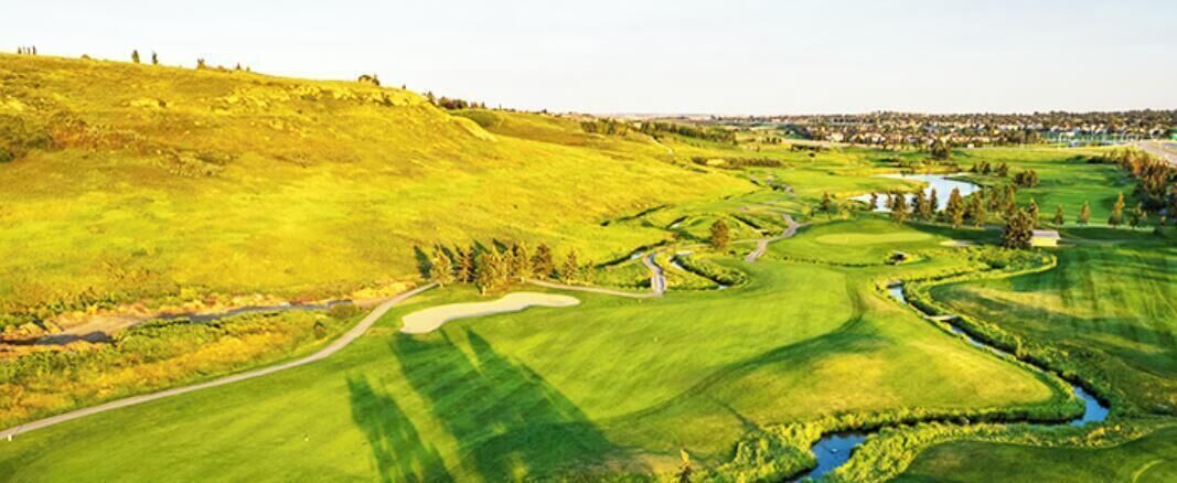 Un campo de golf con un sendero sinuoso, elementos acuáticos y un pueblo lejano.