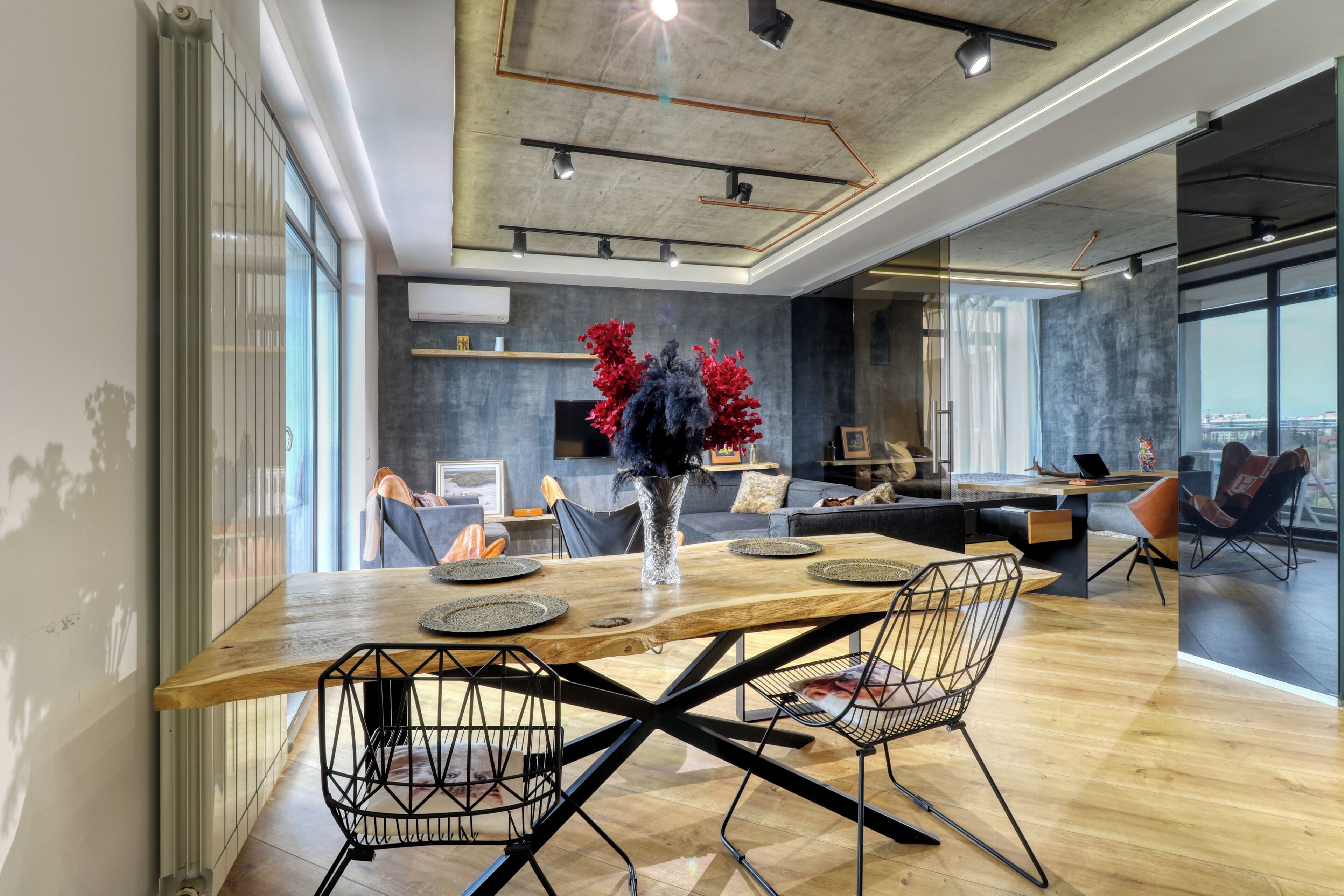 Un espace repas moderne avec une table en bois, des chaises en fil de fer noir et un vase rempli de fleurs rouges.