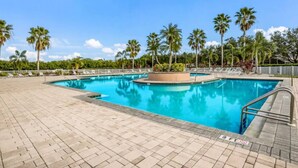 En stor utomhuspool med en kakelbeklädd poolterrass, omgiven av palmer och en klar himmel.