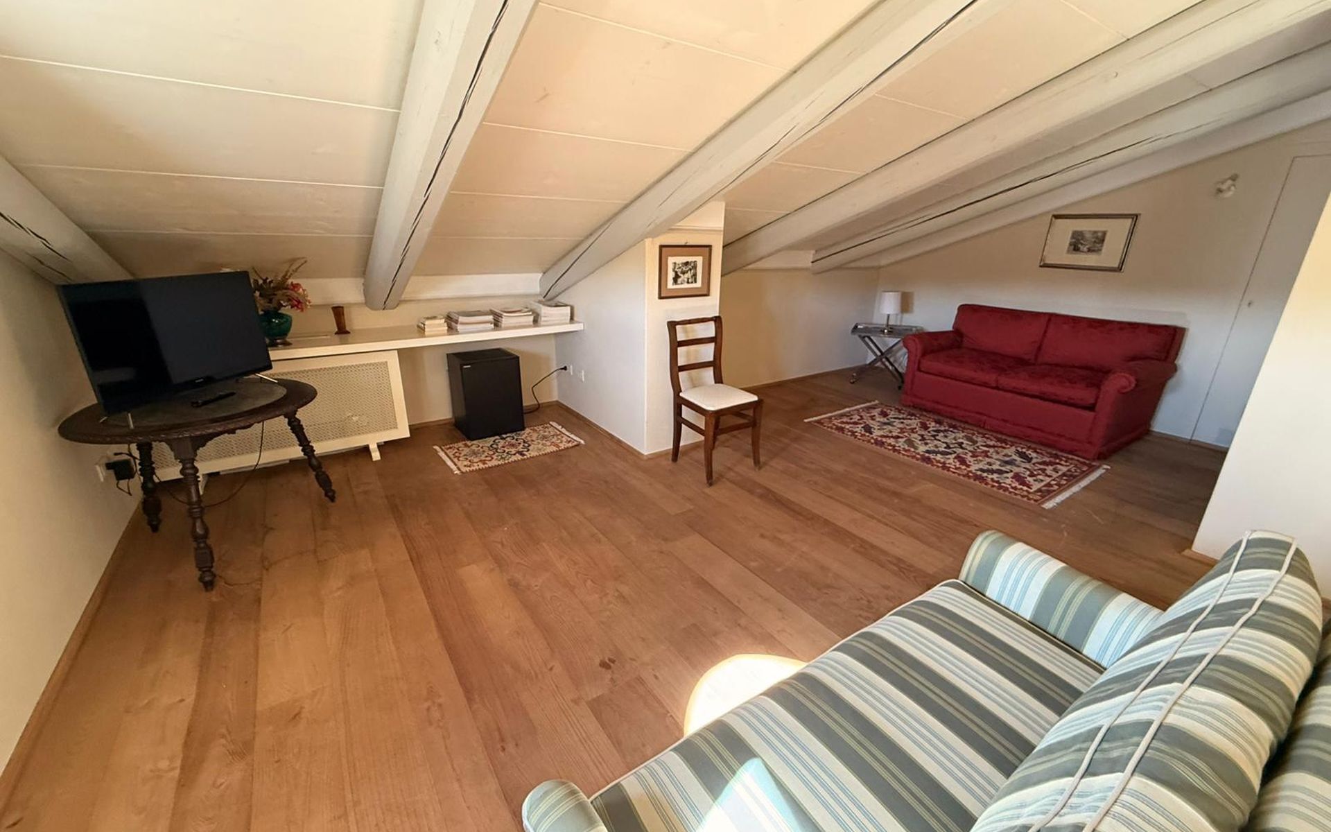 Una sala de estar con televisor, un sofá rojo, un sofá a rayas y piso de madera.