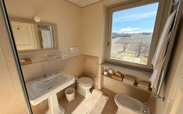 A compact bathroom with a sink, toilet, and bathtub, a window with a view of a field, and a mirror above the sink. - Relais dei Poderi Einaudi (Dogliani)