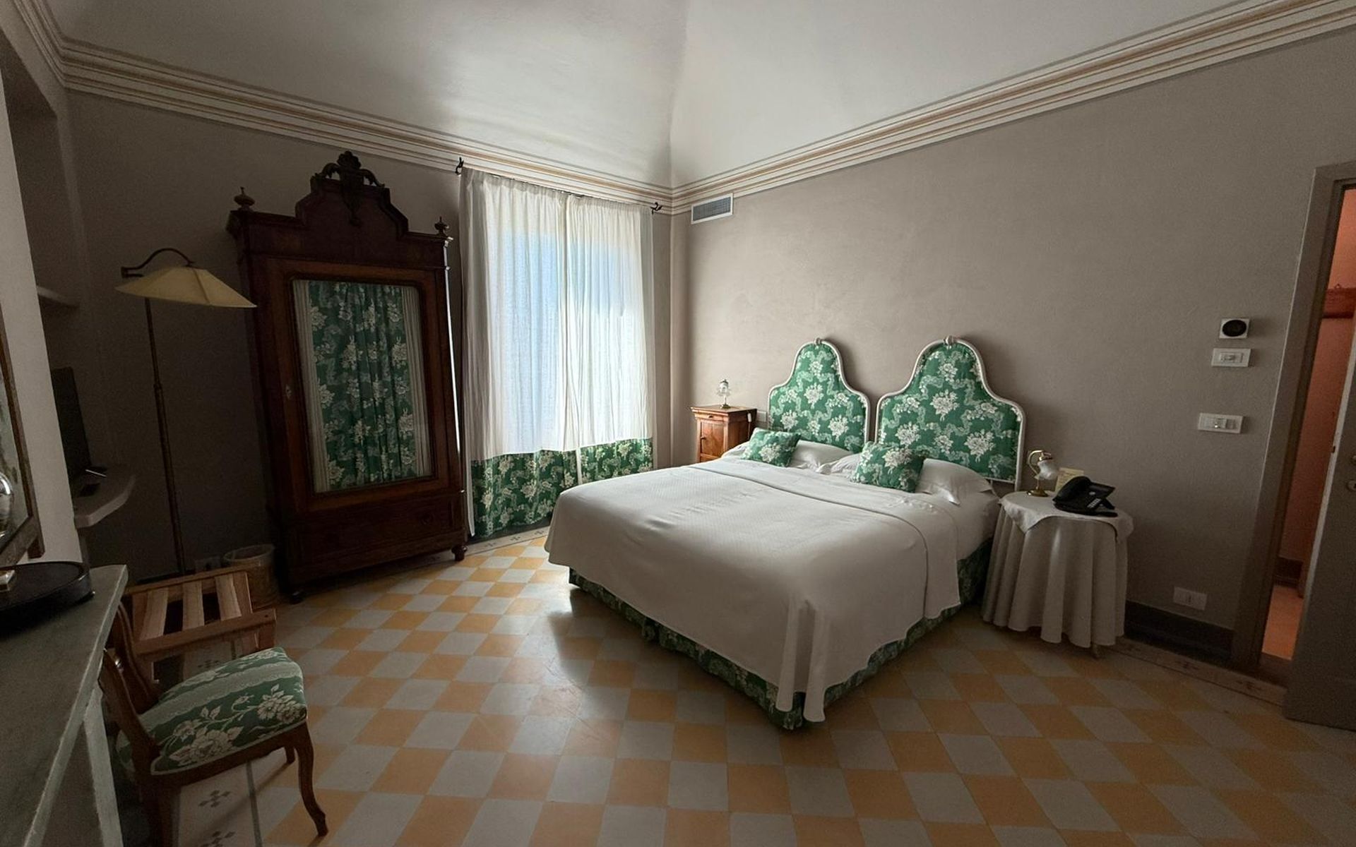 A hotel room with a checkered floor, a bed with floral headboards, a desk with a lamp, and a wardrobe.
