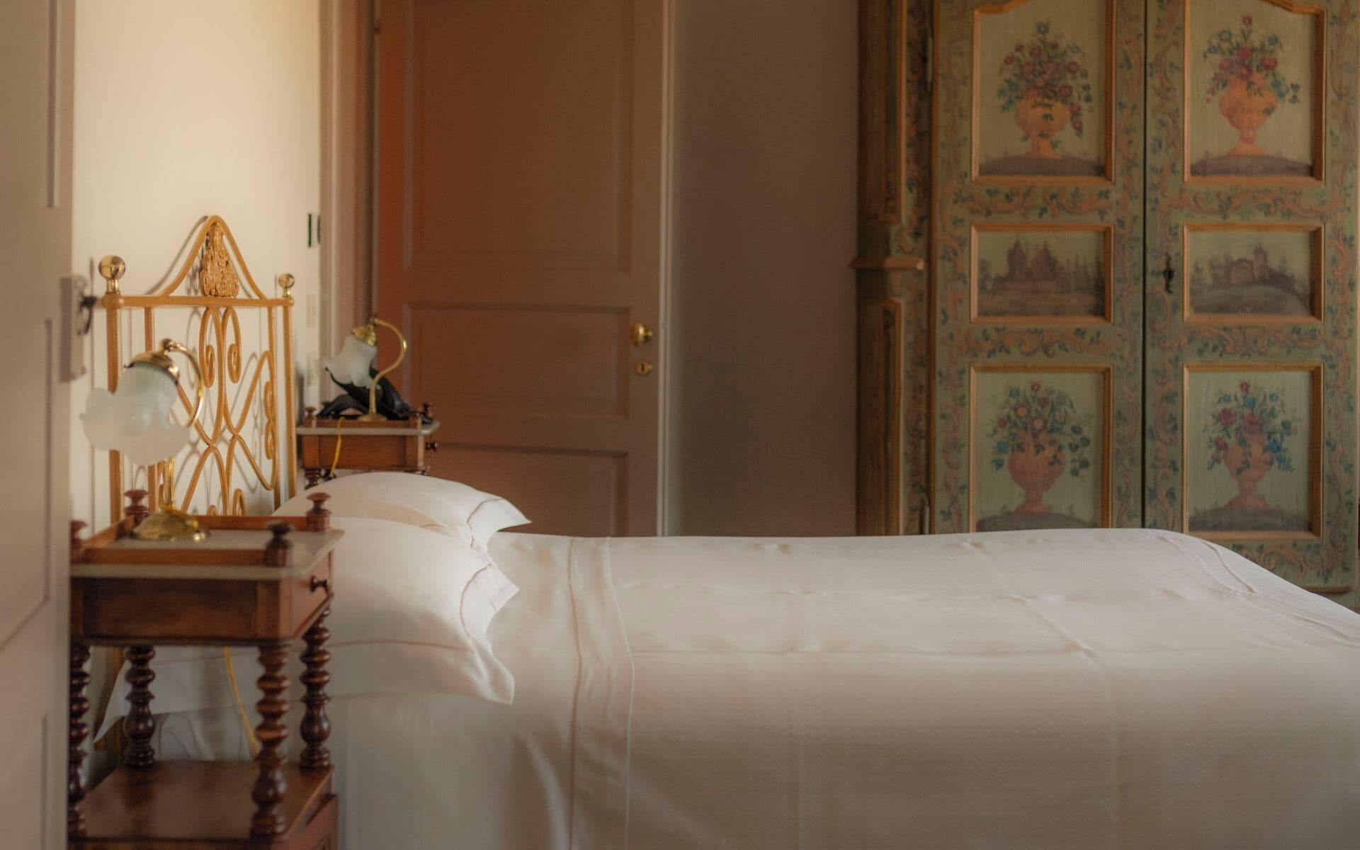 A bedroom with a bed, a nightstand with a lamp, and a door with floral wallpaper.