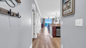 A hallway with wooden flooring, a coat rack, a kitchen area with a view, and a framed picture on the wall. - Tropic Winds 1105 (Panama City Beach)