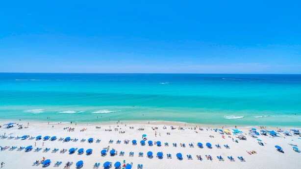 A beach with numerous sun loungers and umbrellas, a clear blue sky, and calm ocean waters. - Tropic Winds 1105 (Panama City Beach)