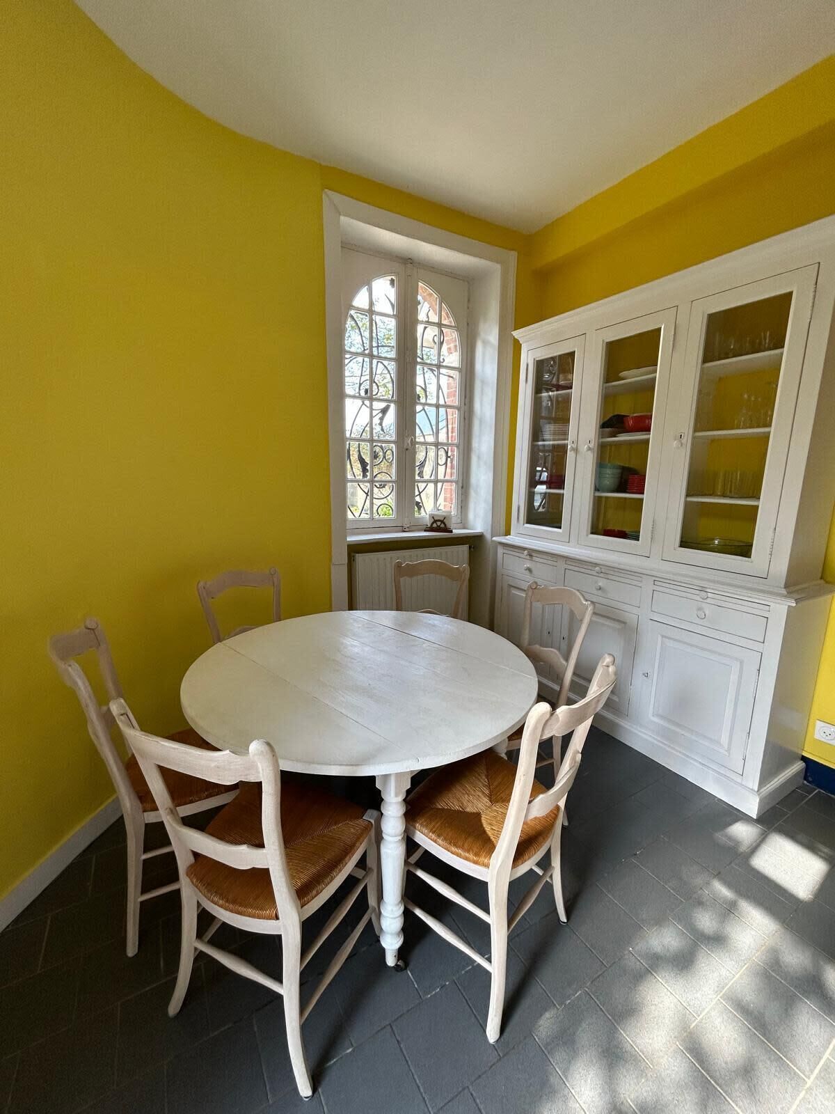 A dining area with a round table and six chairs, a white cabinet with glass doors, and a window with a view of greenery.
