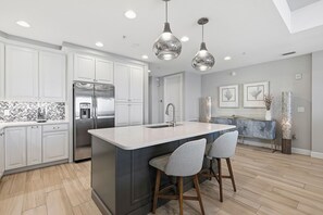 Eine moderne Küche mit weißen Schränken, einem Edelstahl-Kühlschrank, einer zentralen Kochinsel mit Barhocker, Pendelleuchten und einer Küchenwand mit Fischgrätmuster-Fliesen.