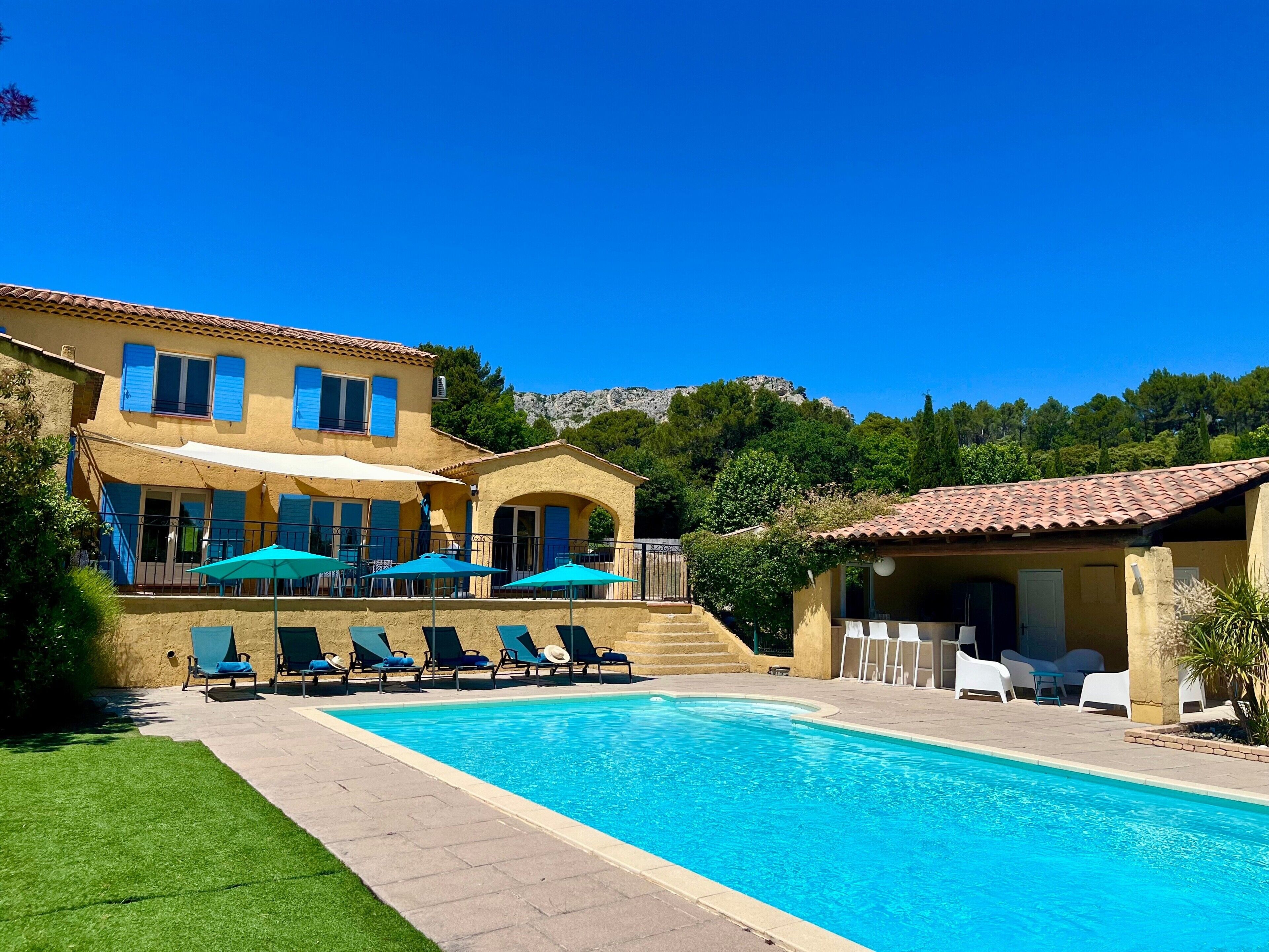 A poolside area with lounge chairs and umbrellas, a pool, and a building with blue shutters.