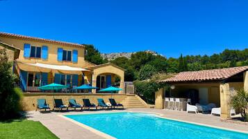A poolside area with lounge chairs and umbrellas, a pool, and a building with blue shutters.