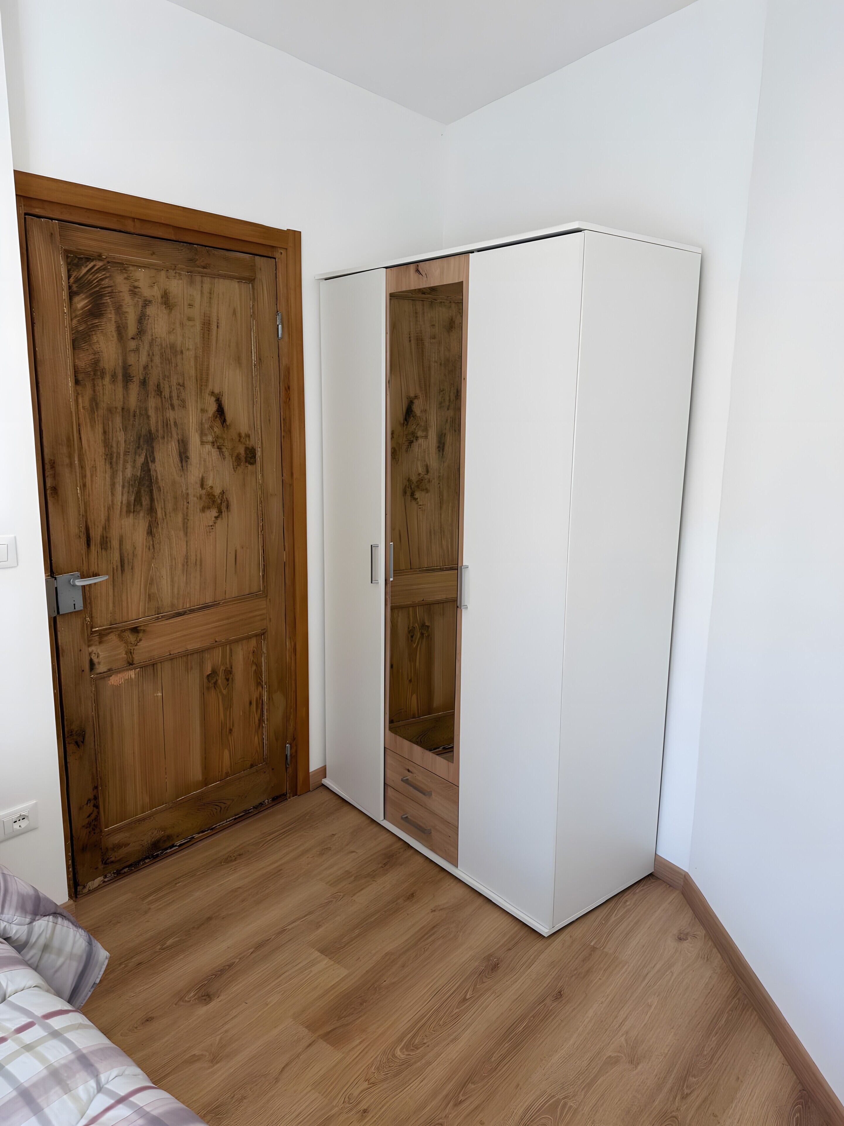 Une pièce avec une porte en bois, une armoire blanche et un lit recouvert d’une couette à carreaux.