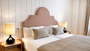 Uma cama de casal com cabeceira estampada, duas luminárias de cabeceira e uma mesinha de cabeceira com abajur.