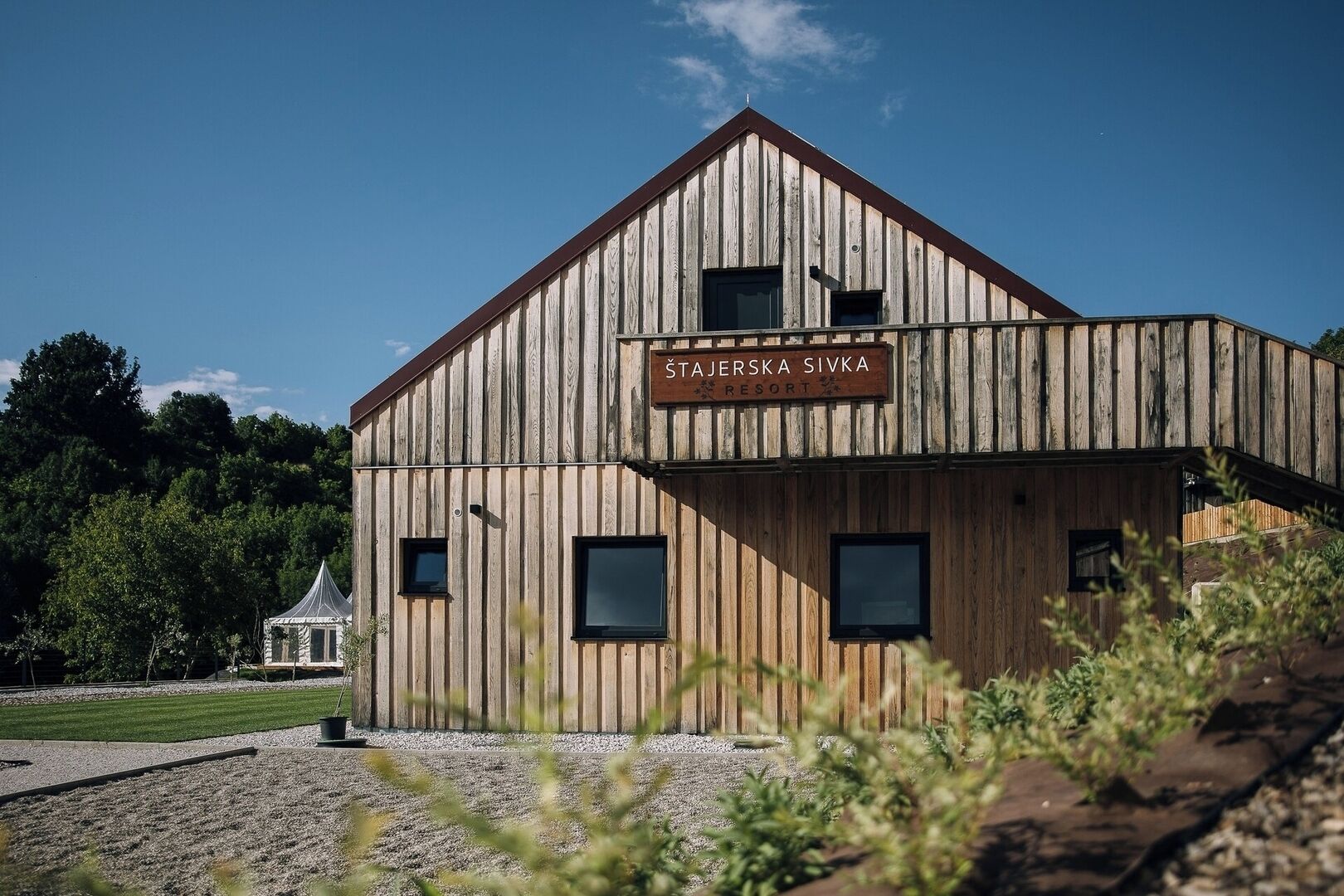 En byggnad av trä med skylten ”Stajerska Sivka Resort”, ett sluttande tak, stora fönster och en grusbelagd mark.