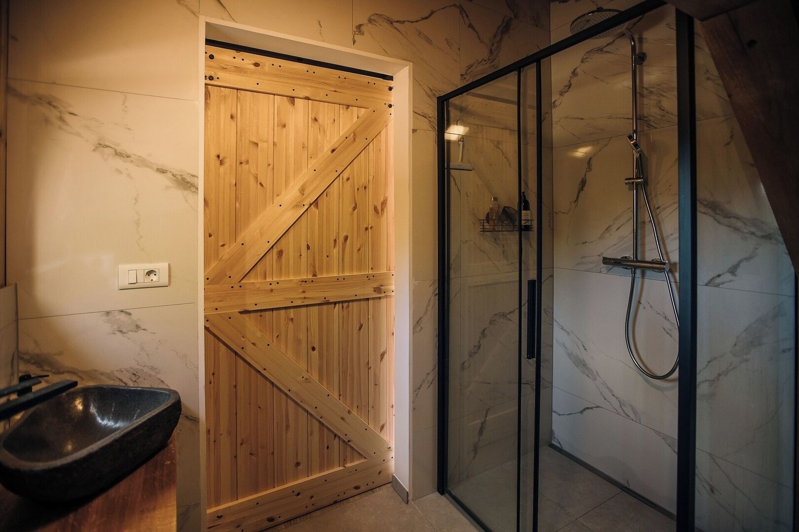 Une porte en bois avec une traverse diagonale, un espace de douche en verre avec un pommeau de douche à main, et un mur recouvert de carreaux de marbre.