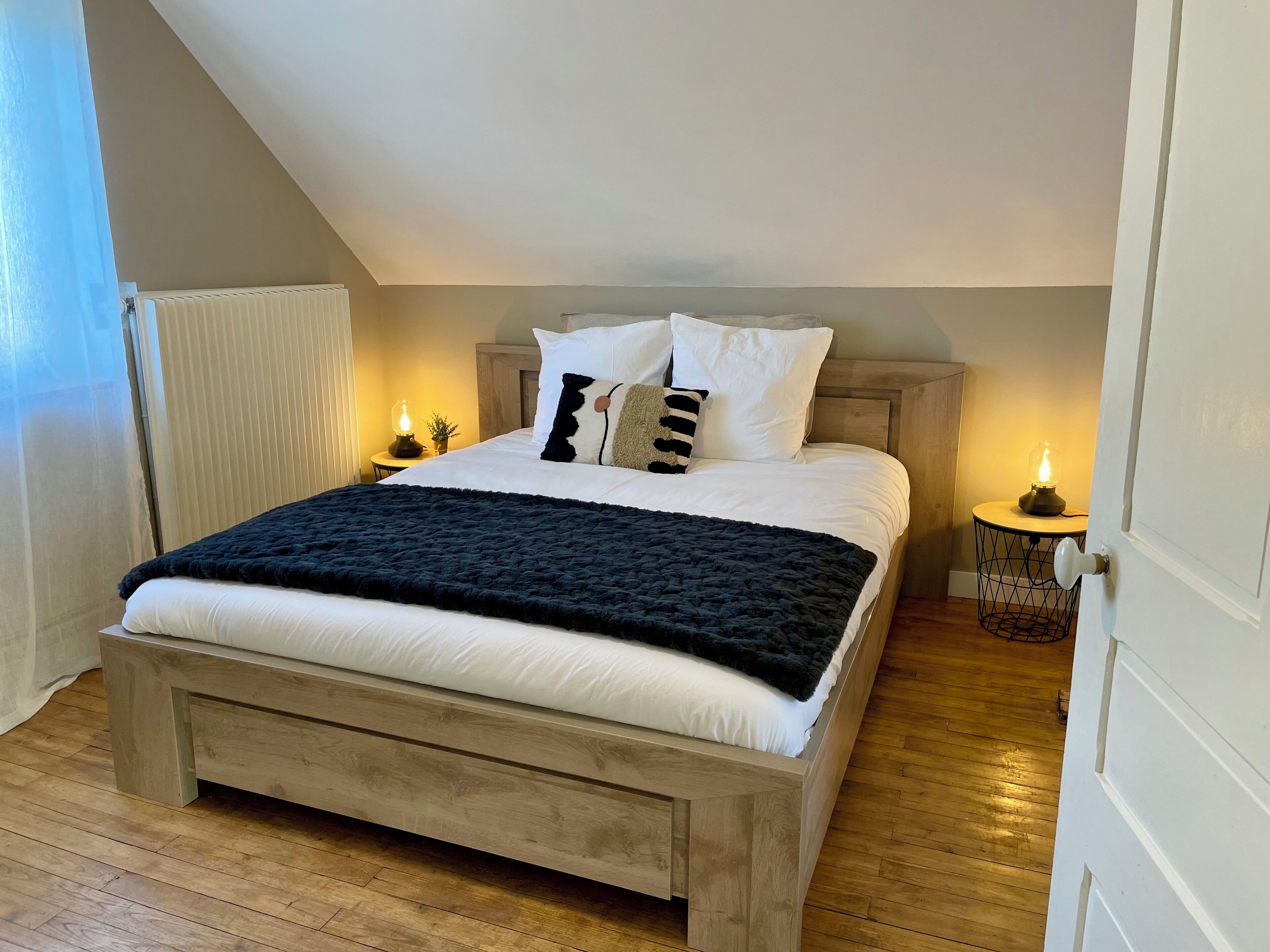 Une chambre à coucher avec un lit en bois, du linge de lit blanc, une couverture foncée et deux lampes de chevet.
