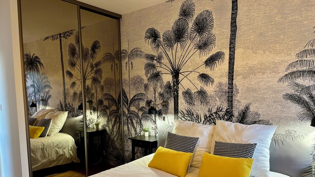 Een slaapkamer met een bed, kussens, een nachtkastje, een lamp en een muur met een tropische plantenmuur.