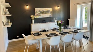 Een eetruimte met een tafel voor zes personen, witte stoelen, een zwarte wand met een stadsgezicht schilderij en een raam met gordijnen.