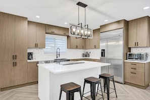 A modern kitchen with wooden cabinets, a stainless steel refrigerator, a built-in island with stools, and a pendant light fixture. - Waterfront Fun! 6BR • Pool • Beach 5 Min (Hollywood)