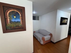 Quarto com piso de madeira, sofá cinza, pintura emoldurada de uma paisagem e uma pequena janela com vista para montanhas.