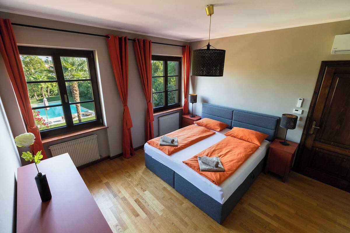 A bedroom with a bed, a bedside table, a lamp, a window with curtains, and a view of a pool.