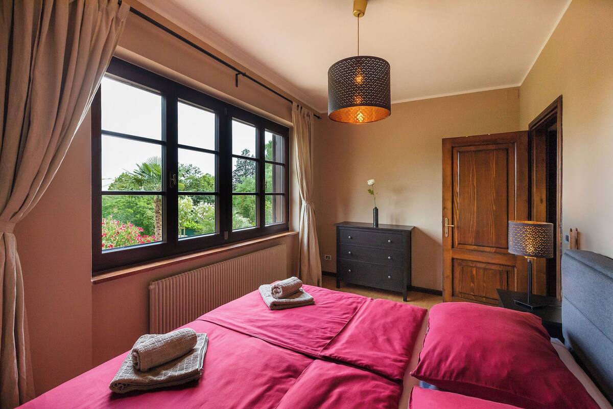 A bedroom with a large window, a bed with pink bedding, a wooden dresser, and a hanging light fixture.