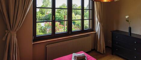 Une chambre avec une grande fenêtre, un radiateur, un lit avec des serviettes, une commode et une lampe suspendue.