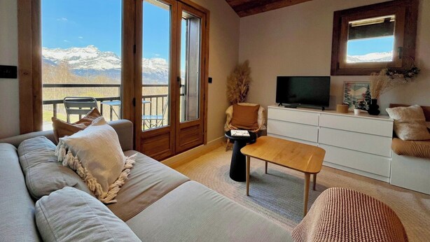 A living room with a sofa, a coffee table, a TV, and a view of mountains through large windows. - Charming studio with mountain view & balcony (Combloux)