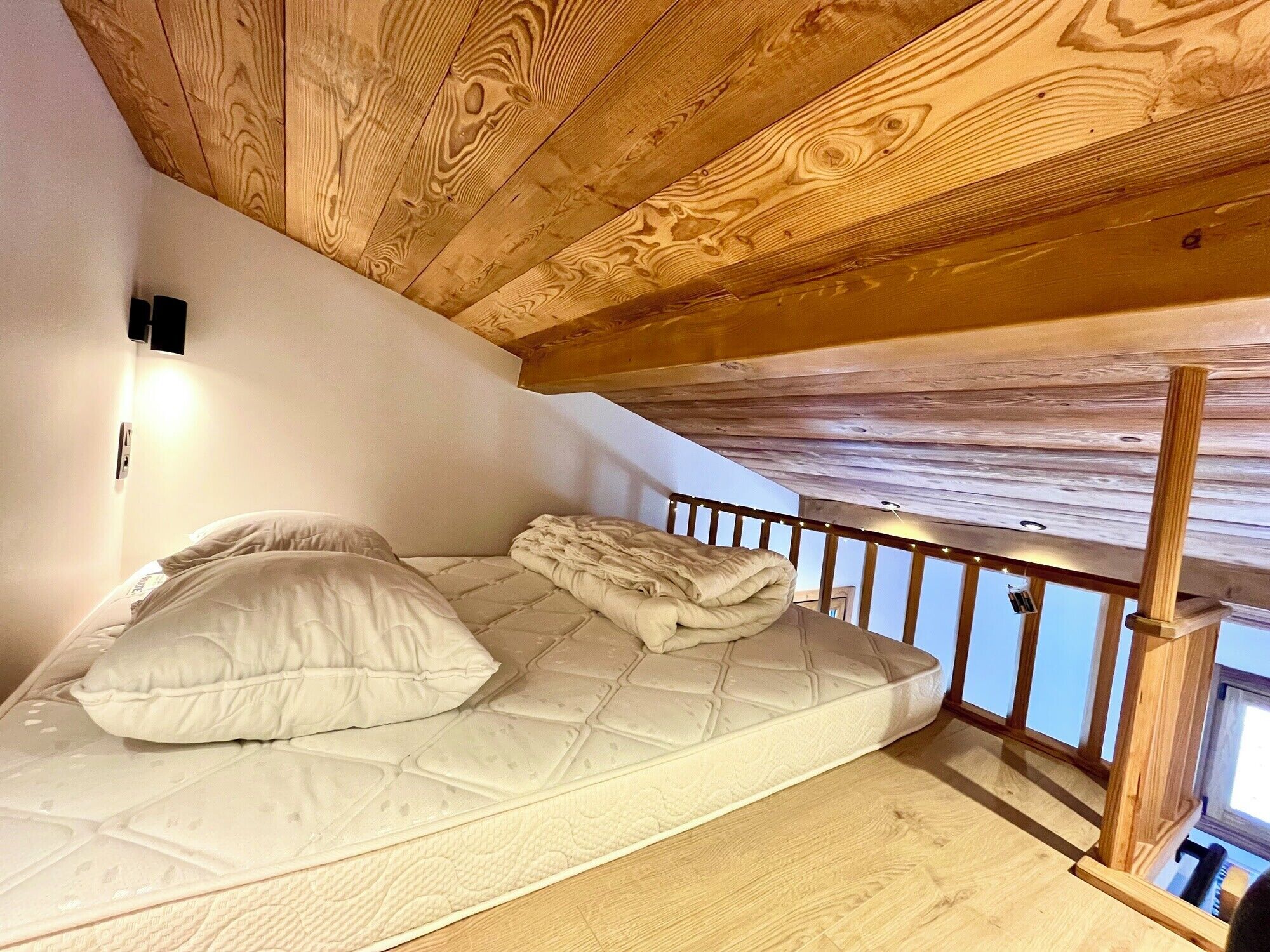 A bunk bed with a mattress and pillows, wooden ceiling beams, a small window, and a wall-mounted light fixture.