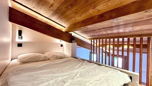A bedroom with a wooden ceiling, a bed with white bedding, and a view through a window with wooden railings. - Charming studio with mountain view & balcony (Combloux)